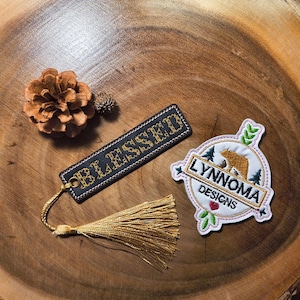 May include: A wooden surface displays a bookmark with "BLESSED" embroidered in gold, a gold tassel, and a patch with a bear and the text "LYNNOMA DESIGNS". A pinecone and small bud are also present.