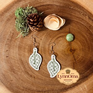 May include: A pair of white leaf-shaped earrings with green embroidery. The earrings are hanging from a wooden surface with a green moss and a pine cone.