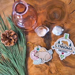 May include: A still life arrangement featuring a clear glass bottle, a pine cone, and a sprig of green pine needles. Two embroidered patches with the text "LYNNOMA DESIGNS" and a capybara design are also present. A small white bear figurine and a seashell are also included.