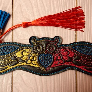 May include: A black leather bookmark with an embroidered owl design. The owl has yellow, red, and blue wings and a blue heart. The bookmark has a blue and red tassel.