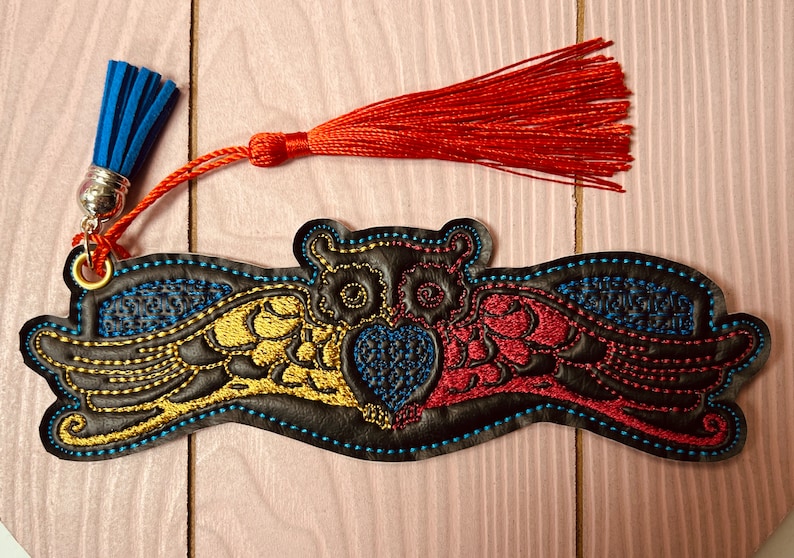 May include: A black leather bookmark with an embroidered owl design. The owl has yellow, red, and blue wings and a blue heart. The bookmark has a blue and red tassel.