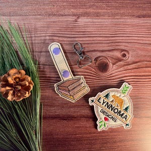 Puede incluir: Un surtido de parches bordados y un llavero sobre una superficie de madera. Un parche presenta un oso y el texto "LYNNOMA DESIGNS". Otro parche representa un sándwich. También hay una piña y vegetación.