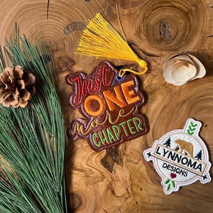 Puede incluir: Un marcapáginas de cuero marrón con el texto "Just One More Chapter" en bordado de colores. Un borla amarilla cuelga de la parte superior del marcapáginas. También se muestra un parche bordado blanco con un oso y el texto "Lynnnoma Designs".
