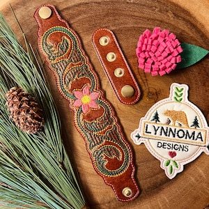 Può includere: Un braccialetto in pelle marrone con un intricato disegno floreale ricamato in verde e rosa. Il braccialetto è abbinato a un piccolo patch in pelle marrone con tre borchie in metallo dorato. Nell'immagine è incluso anche un fiore di feltro rosa con una foglia verde. L'immagine include anche un patch bianco con un orso bruno e il testo "Lynnnoma Designs".