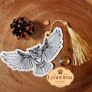 May include: A white embroidered owl bookmark with black stitching and a gold tassel. The bookmark has a gold string attached to the top and the text "LynnOma Designs Est. 2016" is printed on the bottom.