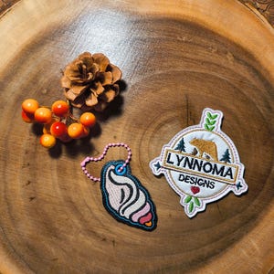 Op de afbeelding: Een assortiment decoratieve items op een houten oppervlak. Inclusief een dennenappel, oranje bessen, een sleutelhanger in de vorm van een zeeschelp en een patch met de tekst "LYNNOMA DESIGNS" en een berenillustratie.