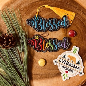 Puede incluir: Dos adornos de fieltro negro con bordado de arco&iacute;ris que dice "Blessed" y un adorno de fieltro blanco con un oso marr&oacute;n y el texto "Lynnnoma Designs".