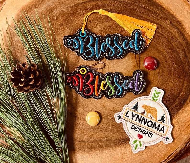 Puede incluir: Dos adornos de fieltro negro con bordado de arco&iacute;ris que dice "Blessed" y un adorno de fieltro blanco con un oso marr&oacute;n y el texto "Lynnnoma Designs".