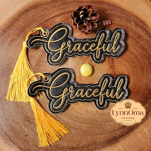 May include: Two black leather bookmarks with the word "Graceful" embroidered in gold script. Each bookmark has a gold tassel attached. The image also includes a pinecone, a yellow bead, and a small logo.
