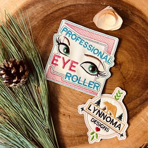 May include: Two embroidered patches. The first patch is white with pink and blue stitching and says "Professional Eye Roller". The second patch is white with pink stitching and says "Lynnnoma Designs" with a bear and flowers.