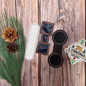 Könnte beinhalten: Eine Auswahl an bestickten Aufnähern und Accessoires. Enthält ein weißes Lesezeichen mit roter Umrandung, marineblaue quadratische Aufnäher, einen Flaschenöffner aus schwarzem Kunstleder und einen Aufnäher mit einem Bären und dem Text "LYNNOMA DESIGNS."