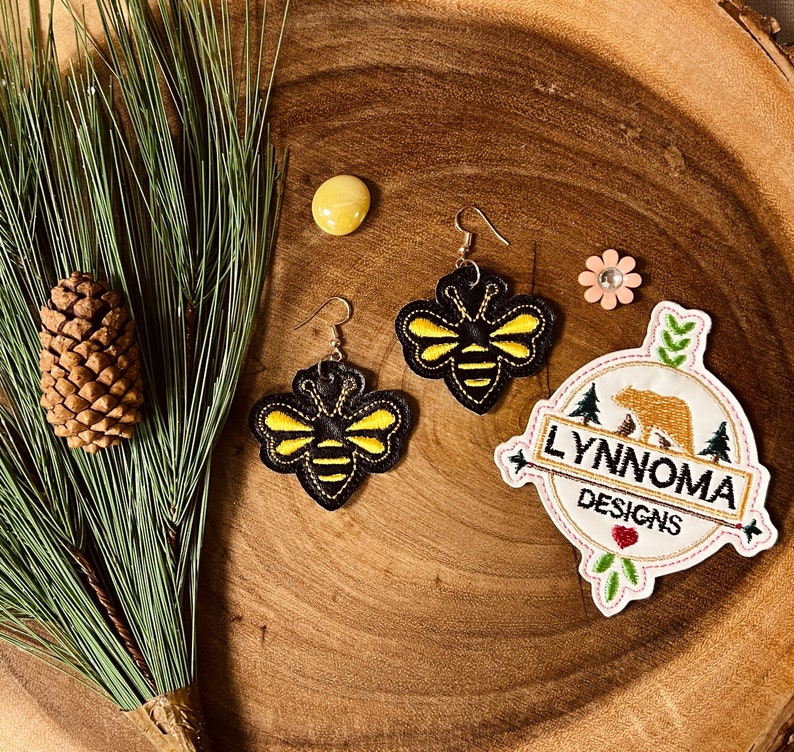 May include: A pair of black and yellow bee earrings with gold accents. The earrings are on a wooden surface with a small pine cone and a green plant. The image also includes a white patch with the text "LYNNOMA DESIGNS" and a brown bear illustration.