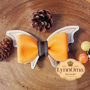 May include: Orange and white bat wing bow with a black center. The bow is on a wooden surface with pine cones and small decorative buttons.