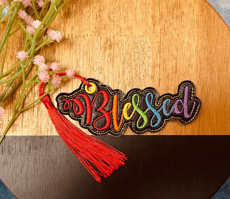 Puede incluir: Un marcap&aacute;ginas de tela negra con la palabra "Blessed" bordada en colores arco&iacute;ris. El marcap&aacute;ginas tiene un borla roja.