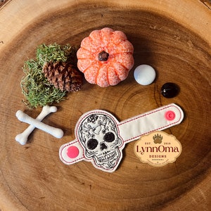 May include: White fabric wrist cuff with a black and white embroidered sugar skull design. The cuff has two pink snap buttons and a white label with the text "Lynn Oma Designs Est. 2016".
