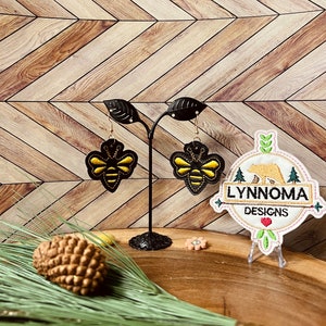 May include: A pair of black leather earrings with gold bee designs. The earrings are displayed on a black stand with a white and brown embroidered logo that says "Lynnnoma Designs".