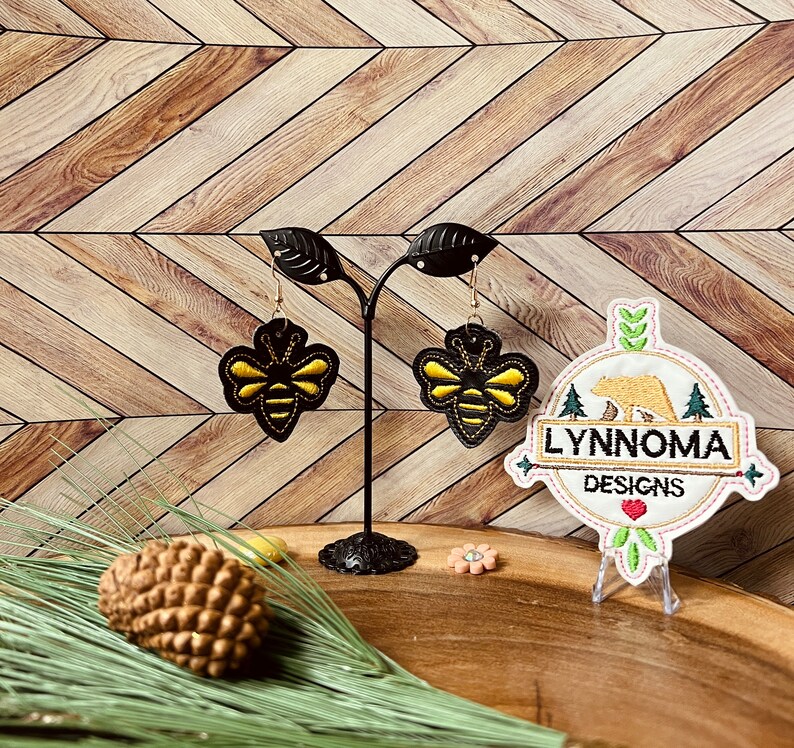 May include: A pair of black leather earrings with gold bee designs. The earrings are displayed on a black stand with a white and brown embroidered logo that says "Lynnnoma Designs".