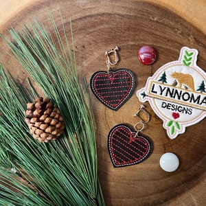Puede incluir: Un par de pendientes en forma de corazón rojo y negro con cierre de clip dorado. Los pendientes se muestran sobre una superficie de madera con una rama de pino y una pequeña piña. También se puede ver un círculo blanco y un círculo rojo en la imagen. La imagen también incluye un parche bordado blanco con el texto "LYNNOMA DESIGNS" y una ilustración de un oso marrón.