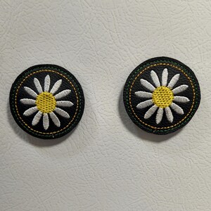 May include: Two round, embroidered patches featuring a daisy design. Each patch has a black background, a white daisy with a yellow center, and a green and yellow stitched border. The patches are likely for clothing or accessories.