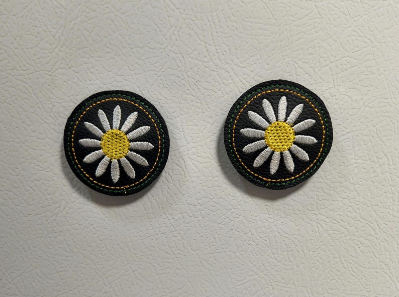 May include: Two round, embroidered patches featuring a daisy design. Each patch has a black background, a white daisy with a yellow center, and a green and yellow stitched border. The patches are likely for clothing or accessories.