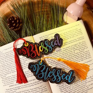 Puede incluir: Dos marcap&aacute;ginas bordados con la palabra "Blessed" en letras de colores. El marcap&aacute;ginas superior tiene letras de arco&iacute;ris con un borla roja. El marcap&aacute;ginas inferior tiene letras negras con un borla dorada.
