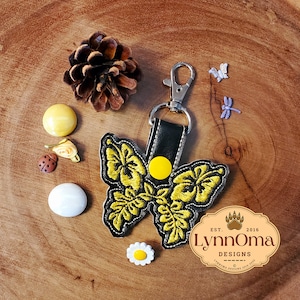 May include: A yellow and black embroidered butterfly keychain with a black leather strap and a yellow button. The keychain is on a wooden surface with a pine cone and other small decorative items.