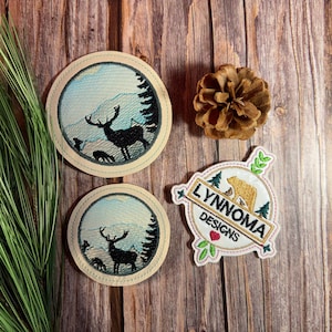 May include: Embroidered patches with nature scenes. Two round patches feature a deer silhouette in a mountain landscape. A third patch has a bear and the text "LYNNOMA DESIGNS". A pine cone and greenery are also present.