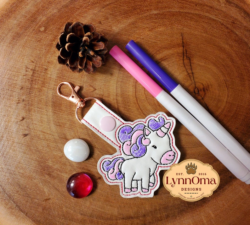 May include: A white unicorn keyring with pink and purple detailing, a gold-coloured clasp, and a white strap. The keyring is next to two markers, a pinecone, and two decorative glass stones. The keyring has the text "LynnOma Designs".
