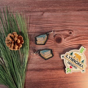 Pode incluir: Brincos e um patch com tema de natureza são exibidos em uma superfície de madeira. Os brincos têm um design azul e amarelo. O patch apresenta um urso, árvores e o texto "LYNNOMA DESIGNS".
