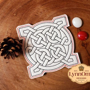May include: A white patch with a black Celtic knot design, outlined with black stitching and a red border. The patch is on a wooden surface, with a pine cone and two decorative buttons. The patch has the text "LynnOma Designs".