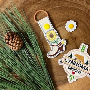 May include: A white leather keychain with a brown leather loop, featuring an embroidered boot with a sunflower and the text "LYNNOMA DESIGNS". The keychain is on a wooden surface with a pine branch and a small pine cone.