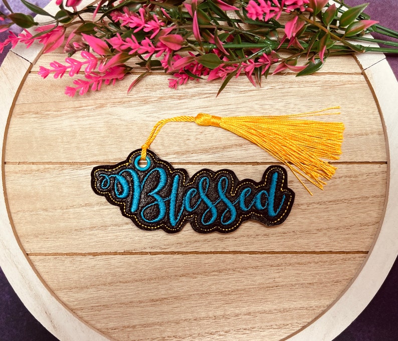 Puede incluir: Un marcap&aacute;ginas de tela negro con bordado azul que dice "Blessed" y un borla amarillo.