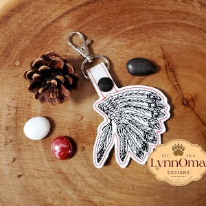 May include: White leather keychain with a black and white embroidered image of a Native American headdress. The keychain has a silver metal clasp and a red stitched border. The text "LynnOma Designs Est. 2016" is printed on the keychain.