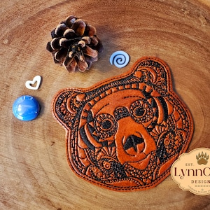 May include: A brown leather bear head patch with intricate black embroidery. The patch is on a wooden surface with a pine cone, a blue stone, and a white heart.