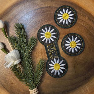 May include: A wooden coaster set with a daisy design. The set includes three round coasters and one elongated coaster, all with a black background, white petals, and yellow centers. A sprig of greenery and white flowers are also present.