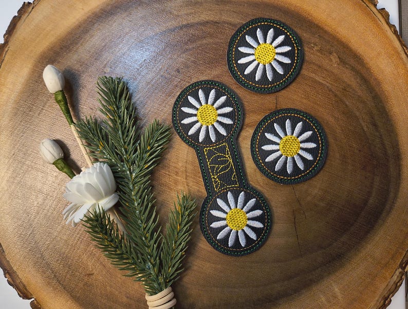 May include: A wooden coaster set with a daisy design. The set includes three round coasters and one elongated coaster, all with a black background, white petals, and yellow centers. A sprig of greenery and white flowers are also present.