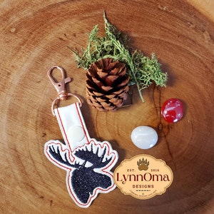 May include: White keychain with a black moose head design and red stitching. The keychain has a rose gold metal ring and a white snap closure. The keychain is on a wooden surface with a pine cone and moss.