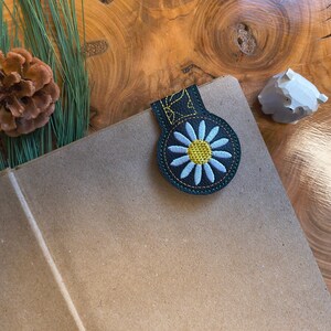 May include: A dark green bookmark with a daisy design, featuring a yellow center and light blue petals, is attached to a brown notebook. A pine cone and greenery are in the background, set on a wooden surface.