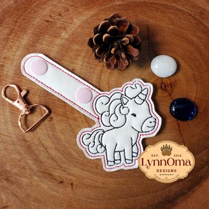May include: A white unicorn keychain with black outline and red stitching. The keychain has a white strap with two pink press studs and a rose gold clasp. The image also includes a pine cone, two white stones, and a dark blue stone.