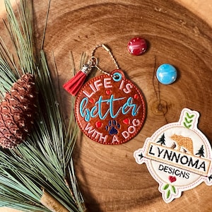 May include: A brown leather keychain with a red tassel and the text "Life is better with a dog" in blue and white embroidery. The keychain also features a paw print and a red heart. The keychain is on a wooden surface with a pine branch and a small blue and red glass bead.