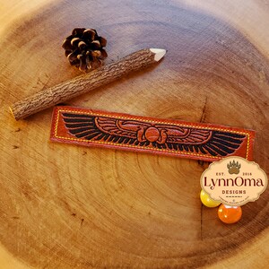 May include: A brown leather bookmark with a black and gold design of a winged scarab beetle. The bookmark has yellow stitching around the edges and the text "Lynn Oma Designs Est. 2016" is visible in the lower right corner.