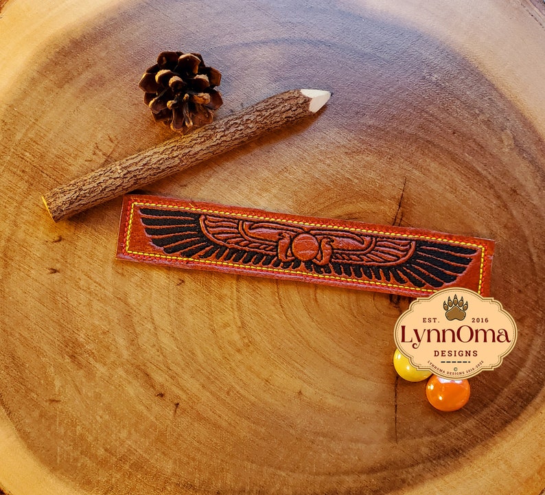 May include: A brown leather bookmark with a black and gold design of a winged scarab beetle. The bookmark has yellow stitching around the edges and the text "Lynn Oma Designs Est. 2016" is visible in the lower right corner.