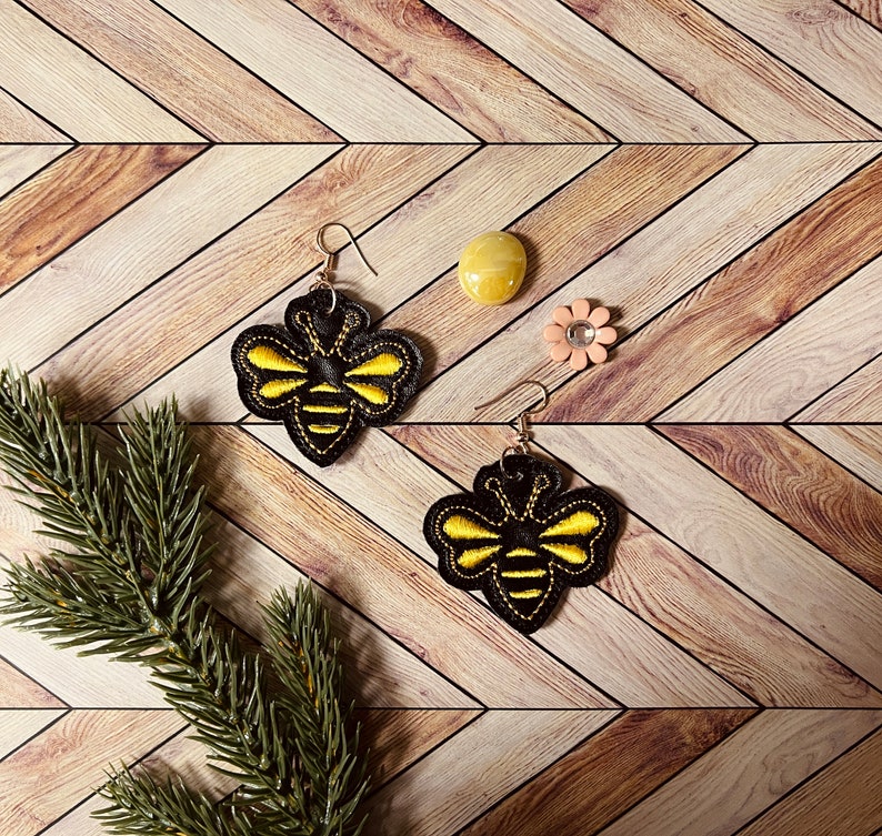 May include: A pair of black and yellow bee-shaped earrings with a woodgrain background.