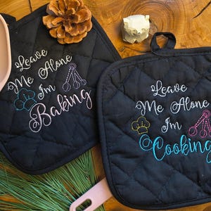 Op de afbeelding: Zwarte, gewatteerde pannenlappen met geborduurde tekst "Leave Me Alone I'm Baking" en "Leave Me Alone I'm Cooking". Een roze siliconen lepel en spatel zijn ook in de afbeelding. Een dennenappel en een klein wit gesneden object zijn ook zichtbaar.
