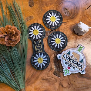 May include: An assortment of embroidered patches and a wooden carving on a wood-grain surface. The patches feature daisy designs and a logo with text that reads "LYNNOMA DESIGNS". A pine cone and pine needles are also present.