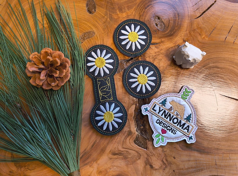 May include: An assortment of embroidered patches and a wooden carving on a wood-grain surface. The patches feature daisy designs and a logo with text that reads "LYNNOMA DESIGNS". A pine cone and pine needles are also present.