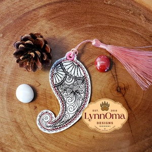 May include: A white bookmark with a black and red paisley design. The bookmark has a pink tassel attached to the top. The bookmark is on a wooden surface with a pine cone and a red stone.