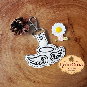 May include: White angel wing keychain with a black outline and a halo. The keychain is attached to a silver split ring. The keychain is on a wooden surface with a pine cone and a white daisy.