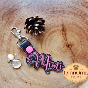 Könnte beinhalten: Ein schwarzer Leder-Schlüsselanhänger mit dem in Pink gestickten Wort "Mimi" und einem rosa Knopf. Der Schlüsselanhänger hat einen silbernen Metallverschluss. Der Schlüsselanhänger liegt auf einer Holzoberfläche mit einer Kiefernzapfen und einer kleinen rosa Blume.