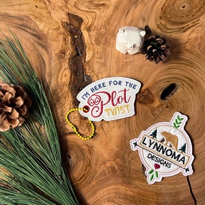 May include: Two embroidered patches with text and designs are displayed on a wooden surface. One patch reads "I'M HERE FOR THE PLOT TWIST" and the other features a bear and the text "LYNNOMA DESIGNS". A small wooden bear, pine cones, and pine needles are also present.